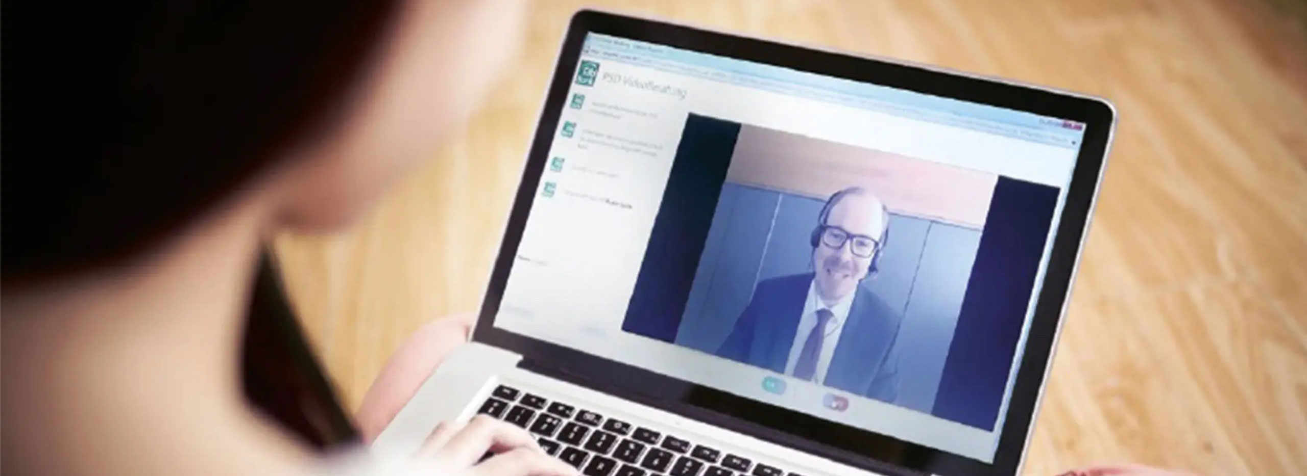 Eine Person führt auf einem Laptop ein Videogespräch mit einem Mann im Anzug und Headset, der auf dem Bildschirm lächelt. Symbolbild für Persönliche Beratung vom Wohnzimmersofa.