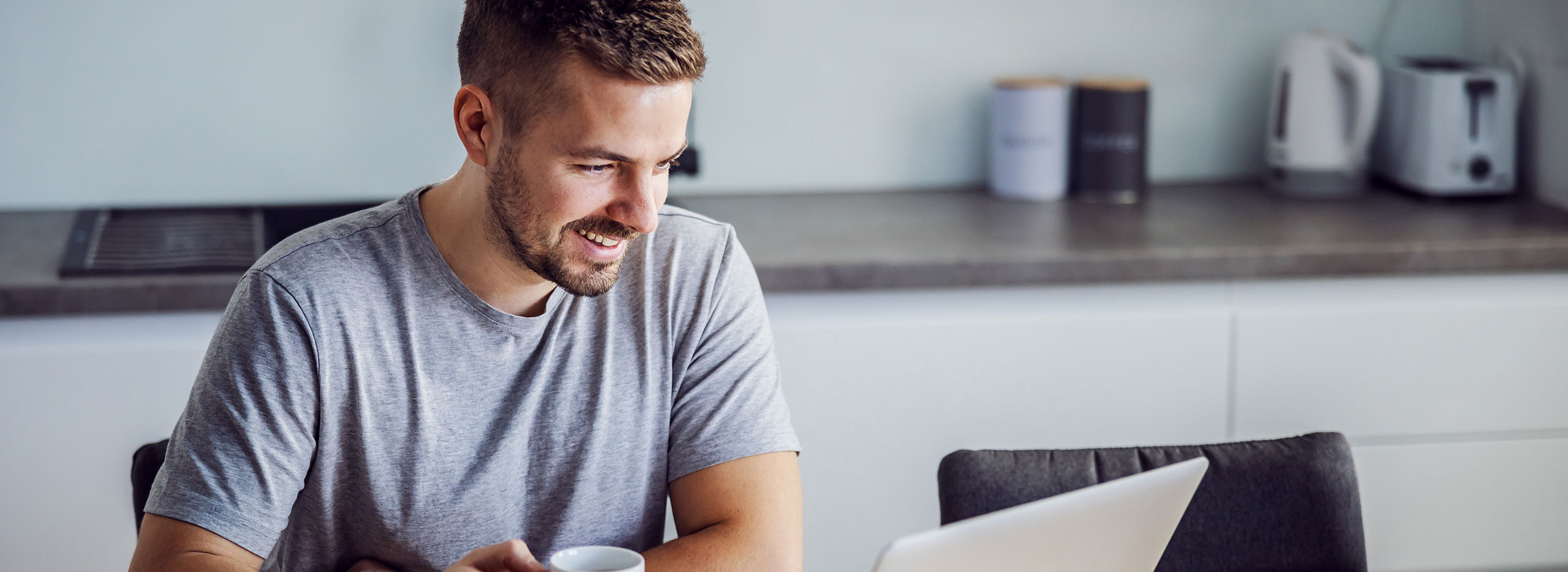 Ein Mann sitzt in einer modernen Küche vor einem Laptop, lächelt und hält eine Tasse in der Hand. Im Hintergrund stehen Küchengeräte wie Wasserkocher und Toaster auf der Arbeitsfläche. Symbolbild für Sicher Surfen im Internet.