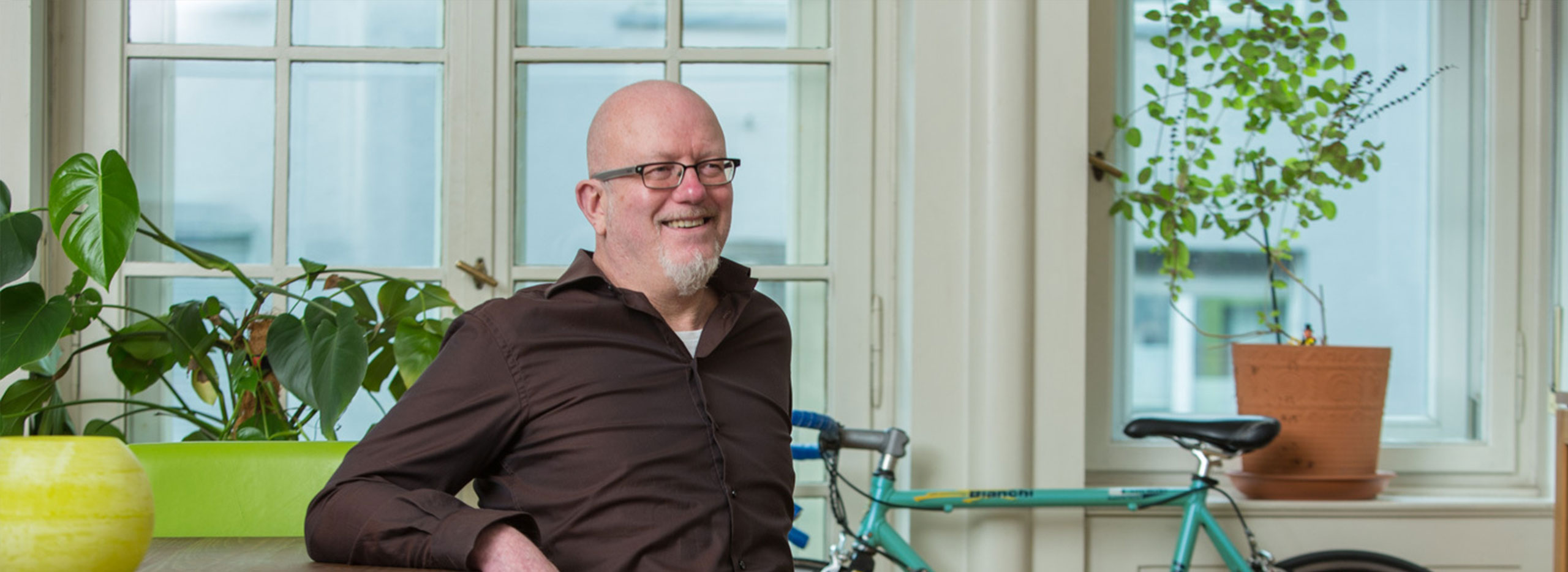 Ein lächelnder älterer Mann mit Brille, Glatze und weißem Kinnbart sitzt entspannt vor einem großen Fenster. Hinter ihm stehen mehrere grüne Zimmerpflanzen und ein türkisfarbenes Fahrrad.
