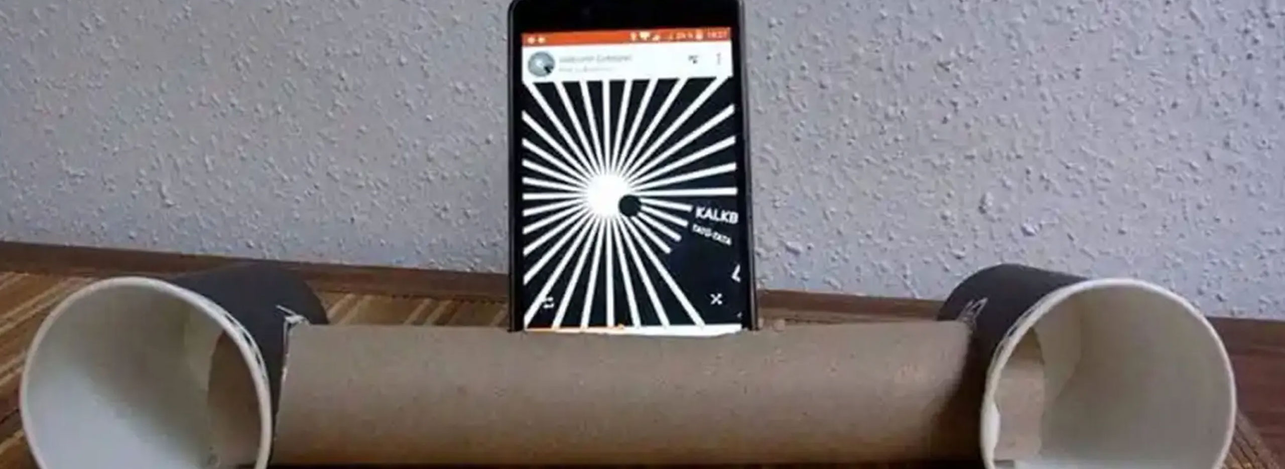 Ein Smartphone steckt in einem Schlitz einer Papprolle; an beiden Enden der Rolle sind Pappbecher befestigt, sodass eine einfache DIY-Handy-Lautsprecherhalter entsteht.