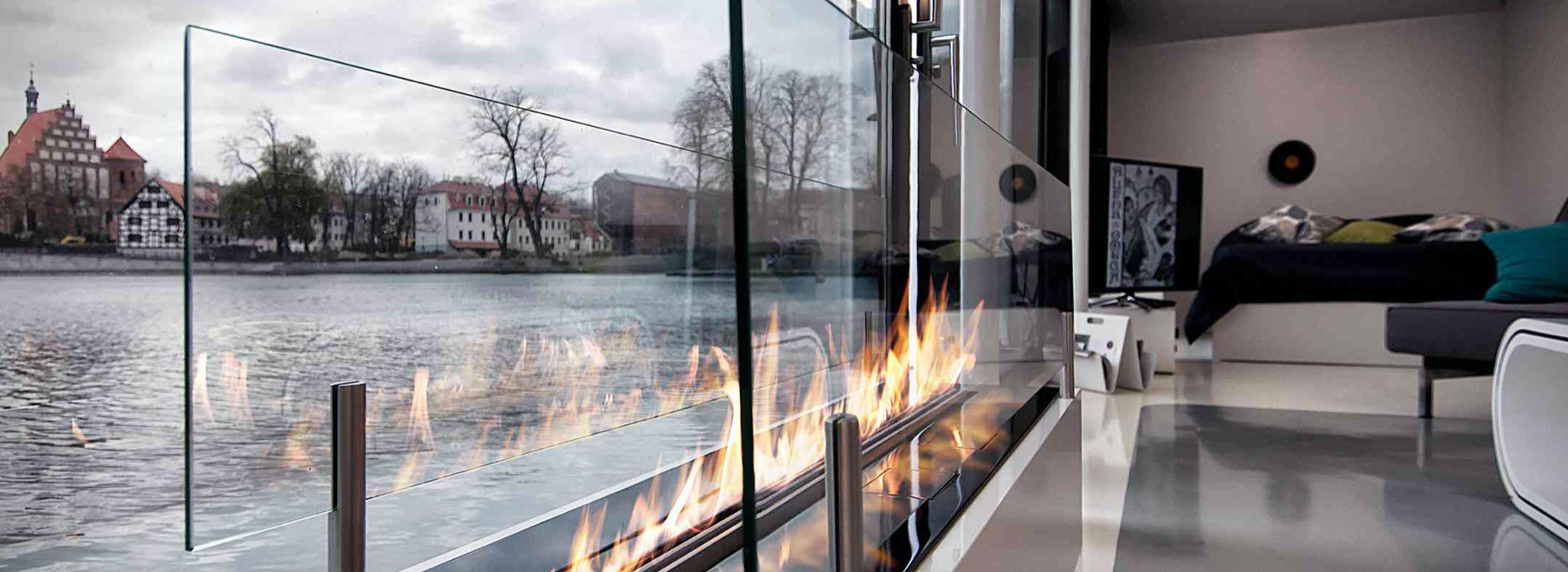 Moderner Wohnraum mit langem, verglastem Kamin, in dem offene Flammen brennen; durch große Fenster ist ein Fluss mit Gebäuden am Ufer zu sehen. Symbolbild für das Thema Kamin und Kaminalternativen.