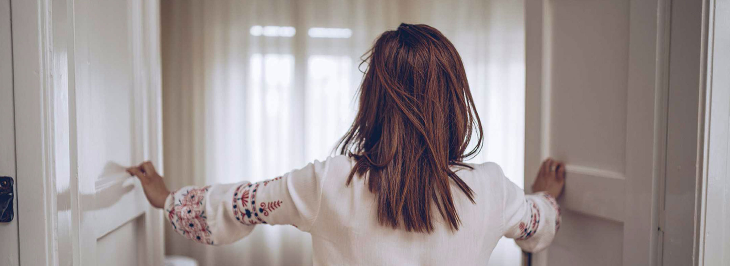 Frau öffnet weit eine Tür zu einem hellen Raum mit weichen Vorhängen, um frische Luft hereinzulassen. Symbolbild für das Thema Raumklima.