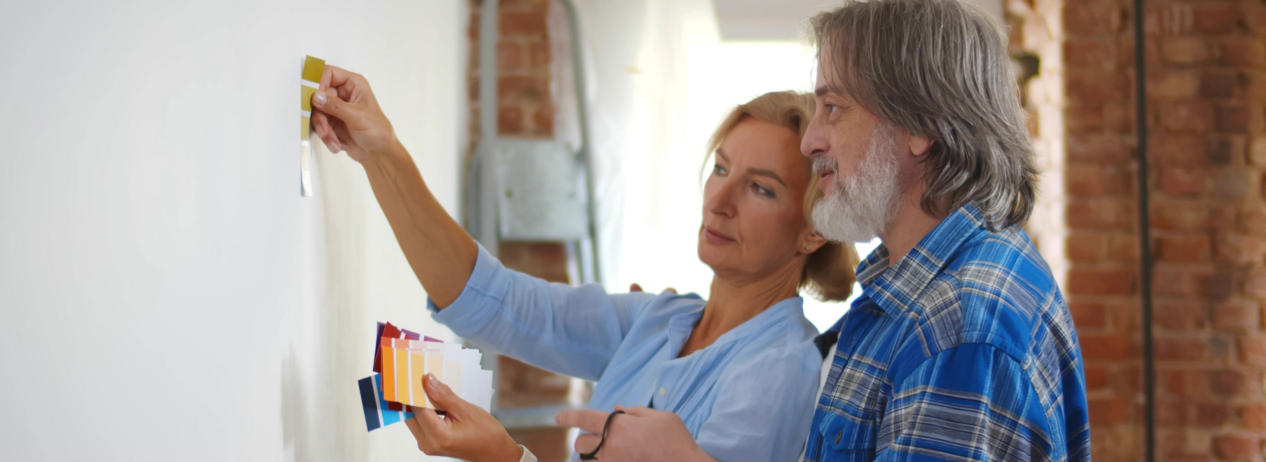 Frau und Mann betrachten gemeinsam Farbmuster an einer weißen Wand, um eine passende Wandfarbe auszuwählen. Symbolbild für Renovieren sobald die Kinder ausgezogen sind.