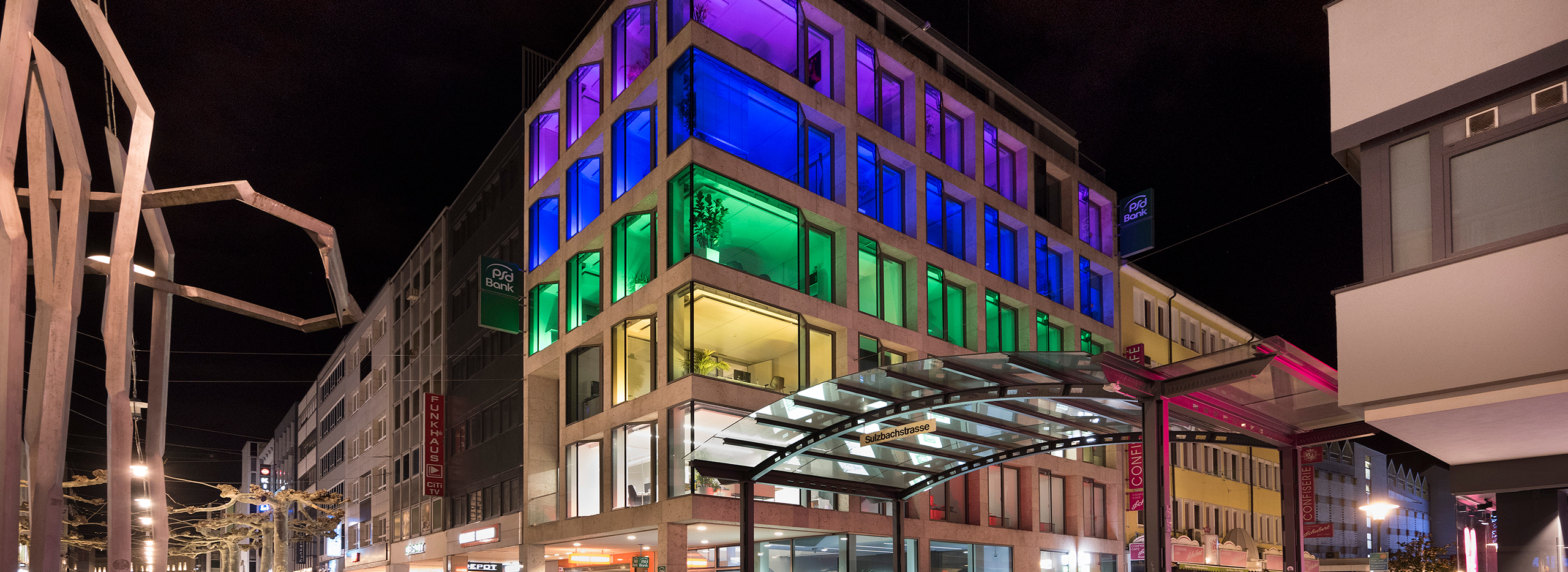Beleuchtetes Geschäftsstelle in Saarbrücken bei Nacht, dessen Fenster in bunten Farben wie Blau, Grün und Pink strahlen; die farbige Illumination setzt ein sichtbares Zeichen für den Frieden.