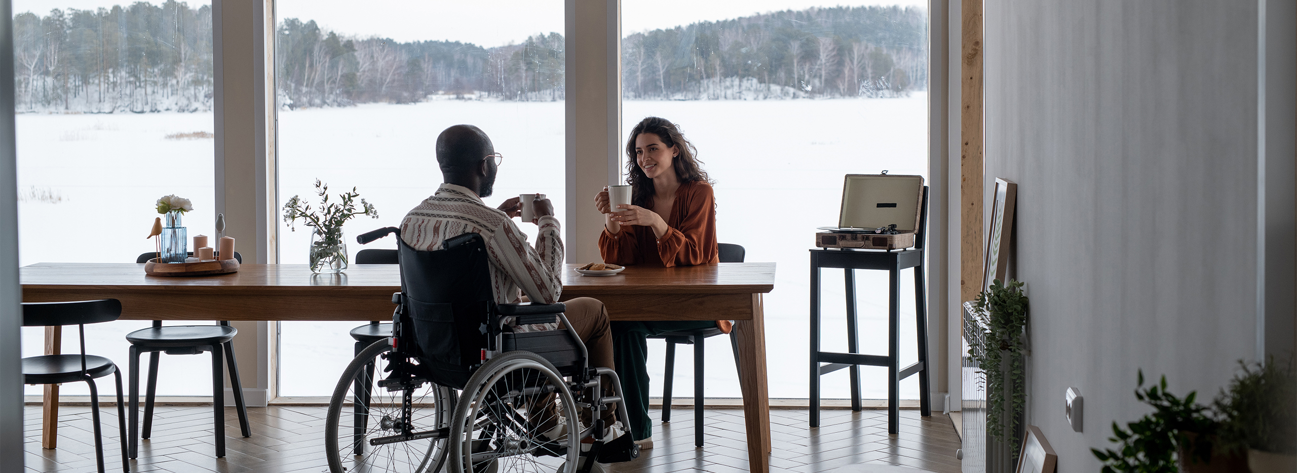 Zwei Personen sitzen einander an einem großen Holztisch gegenüber und trinken Kaffee. Eine Person nutzt einen Rollstuhl. Durch die großen Fenster ist eine verschneite Landschaft zu sehen. Die Szene wirkt ruhig und gemütlich. Symbolbild für das Thema Barrierefreies Wohnen.