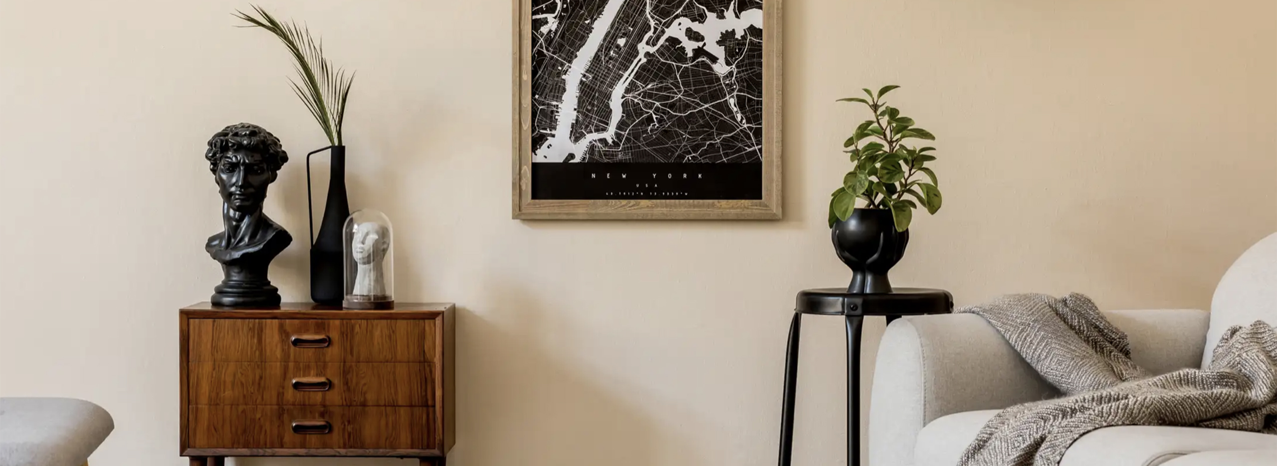 Ein modern eingerichtetes Wohnzimmer mit hellen, natürlichen Farben. Eine kleine Kommode aus Holz steht links, darauf eine dunkle Büste und ein schwarzes Deko-Gefäß mit Halmzweigen. Rechts daneben hängen ein Stadtplan-Poster und eine schwarze Pflanzenvase auf einem schmalen Hocker. Im Vordergrund steht ein helles Sofa mit einer Decke. Symbolbild für das Thema Japandi.