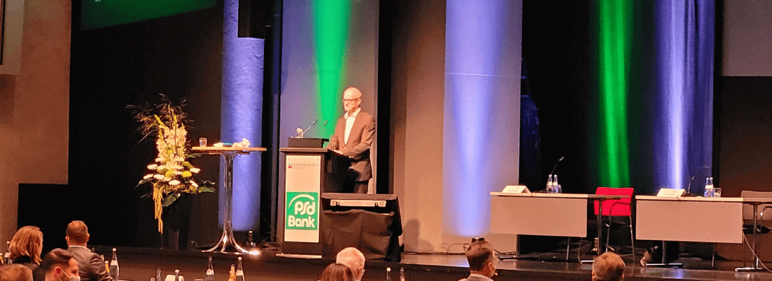 Menschen sitzen in einem großen Saal und hören einem Redner am Podium zu; Bühne mit bunten Lichtsäulen und PSD-Bank-Logo. Symbolbild für das Thema PSD Bank Vertreterversammlung.