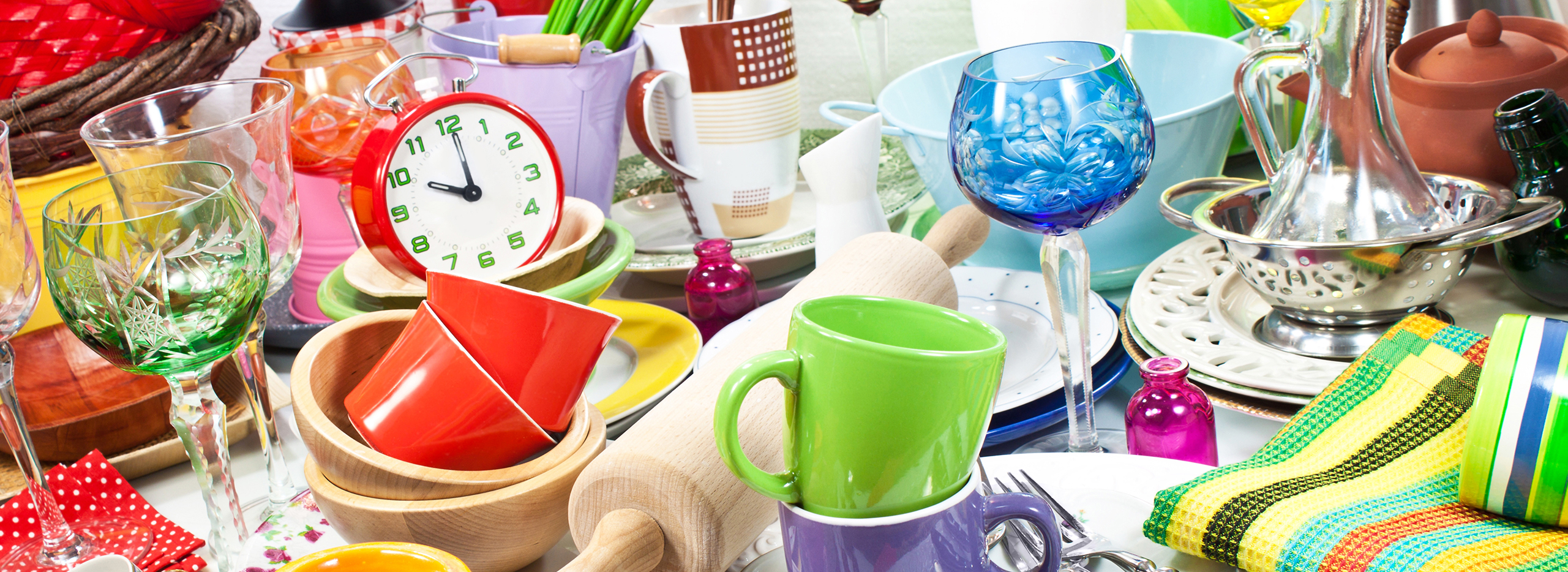 Das Bild zeigt eine bunte Sammlung von Haushaltsgegenständen und Geschirr. Auf einem Tisch stehen verschiedene farbenfrohe Tassen, Gläser, Teller, Schüsseln und Küchenutensilien, darunter ein Nudelholz, eine rote Uhr und bunte Stoffservietten. Symbolbild für Dinge die Ausgemistet werden sollten.
