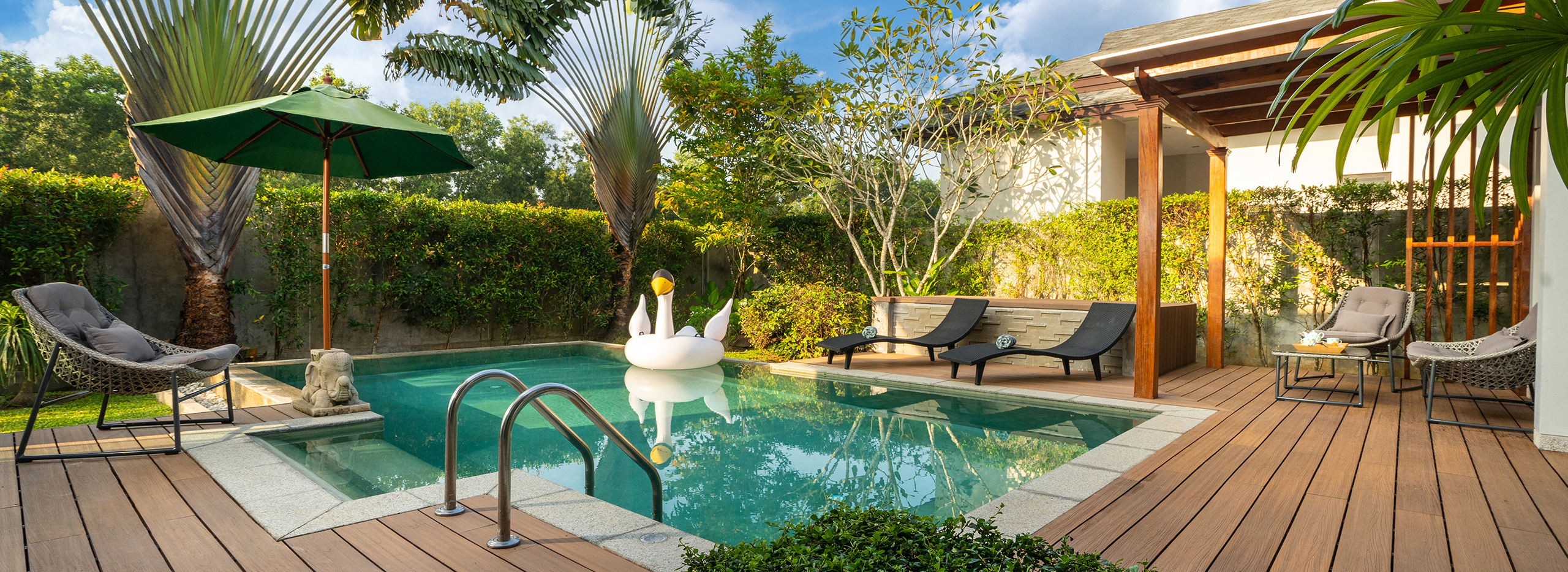 Ein moderner Garten mit Pool, Sonnenschirm und Liegestühlen. Auf dem Wasser schwimmt eine große, weiße Schwanen-Luftmatratze. Rund um den Pool stehen Pflanzen und Palmen. Symbolbild: Der Garten Wellness Tempel.