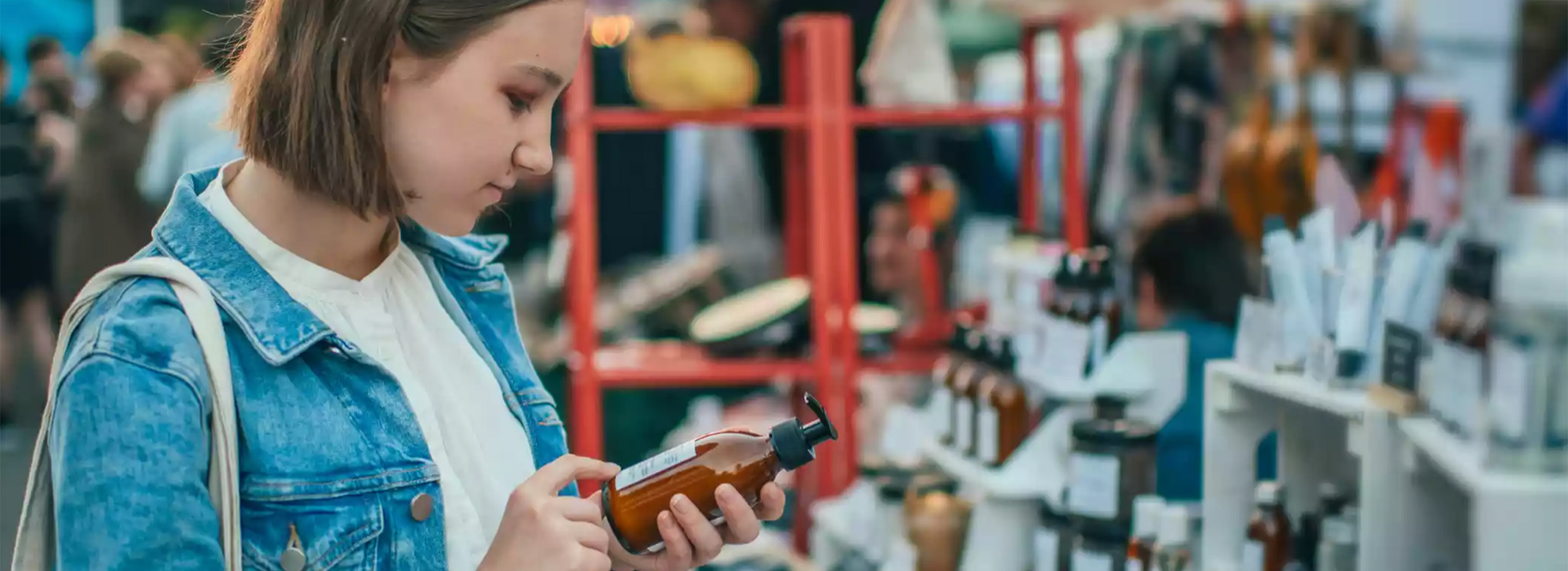 Das Bild zeigt eine junge Frau auf einem Markt oder Basar. Sie trägt eine Jeansjacke und betrachtet aufmerksam eine braune Flasche mit Spender, vermutlich ein Pflege- oder Kosmetikprodukt. Symbolbild für Nachhaltig leben.
