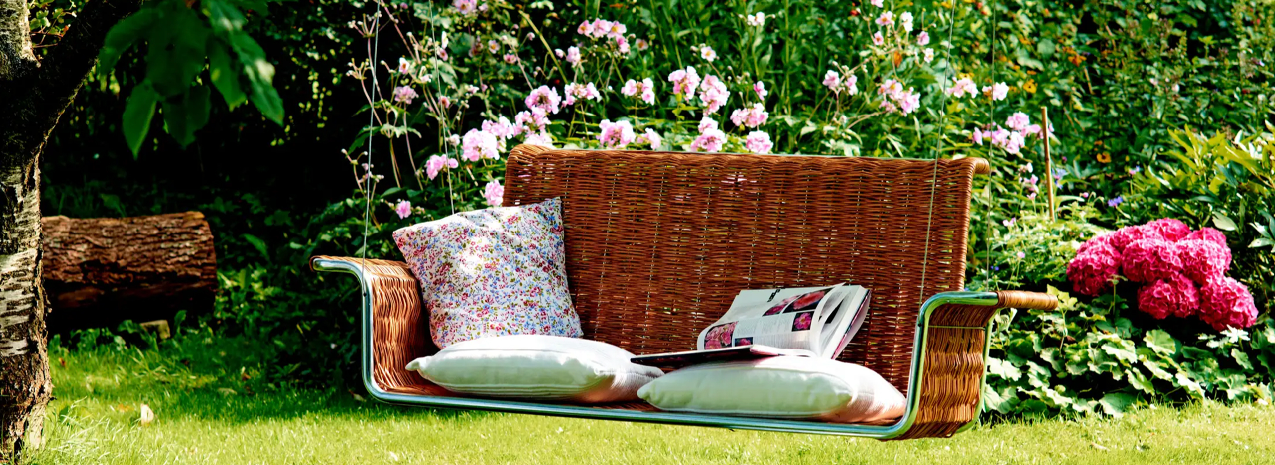Eine geflochtene Gartenschaukelbank mit Kissen hängt unter einem Baum, umgeben von blühenden Pflanzen und sattem Grün – ein gemütlicher Sitzplatz im Garten. Symbolbild für das Thema Gartenmöbel Trends.