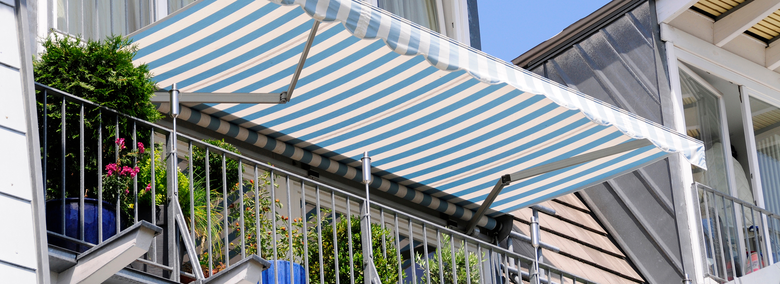 Balkon mit blauer und weiß gestreifter Markise, bepflanzt mit grünen Sträuchern und Blumen in bunten Töpfen, geschützt durch ein Metallgeländer unter blauem Himmel.