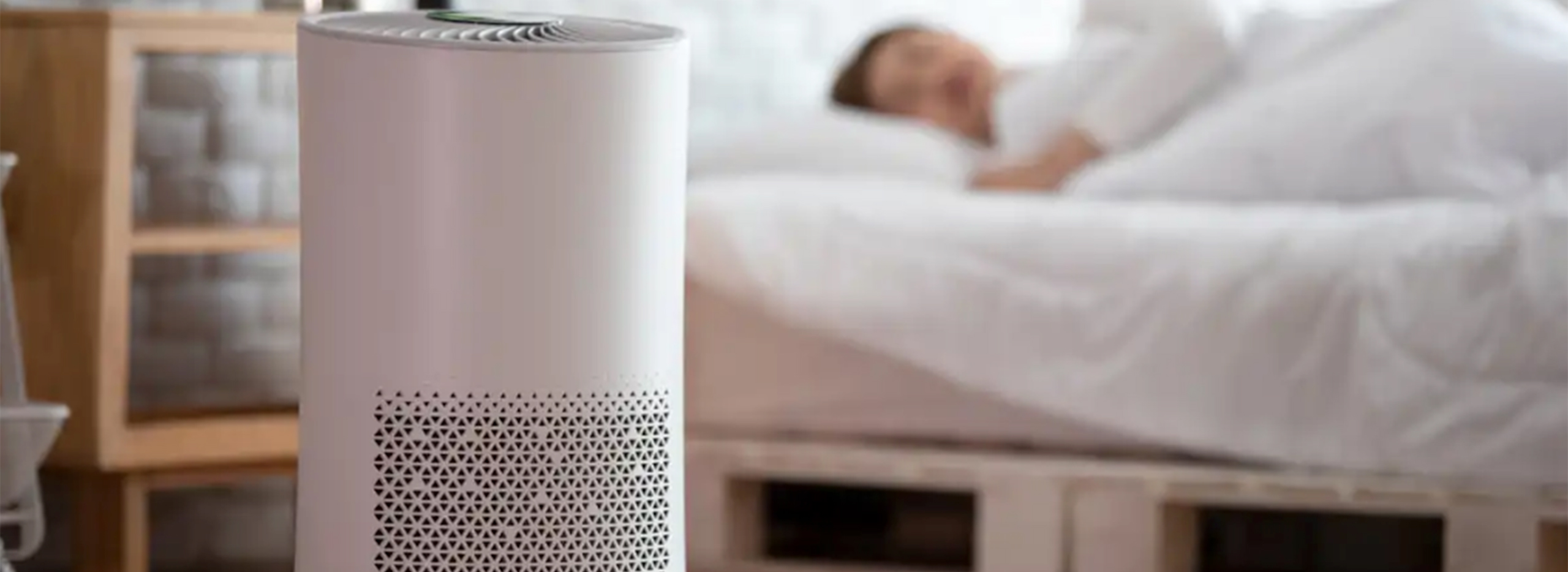 Ein weißer Luftreiniger steht im Vordergrund eines Schlafzimmers auf dem Boden. Er hat ein rundes, modernes Design. Symbolbild für Luftreiniger im Test.