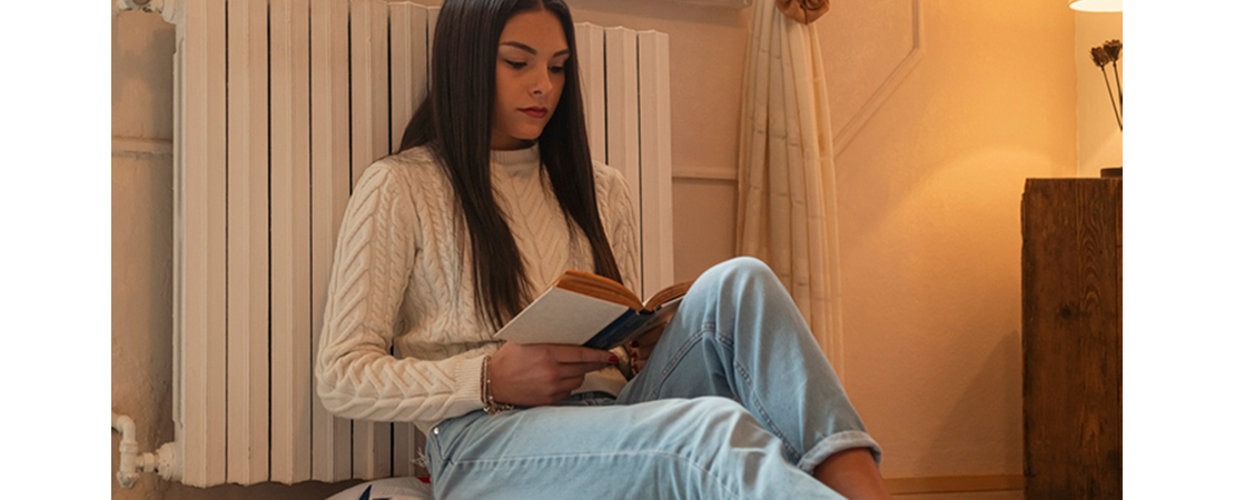Frau in gemütlicher Winterkleidung liest entspannt ein Buch vor einer warmen Heizung im Wohnzimmer.