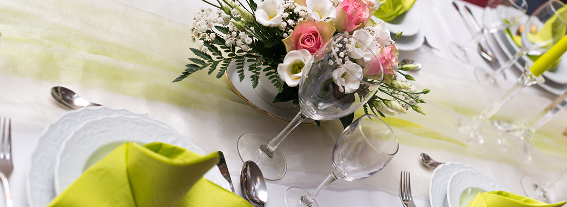 Festlich gedeckter Tisch mit eleganter Blumendekoration aus Rosen, grünen Servietten, weißen Tellern und edlen Weingläsern.