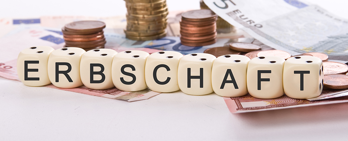 Würfel mit dem Schriftzug „ERBSCHAFT“ stehen auf Euro-Geldscheinen und Münzen, im Hintergrund sind gestapelte Münzen und Geldscheine zu sehen.