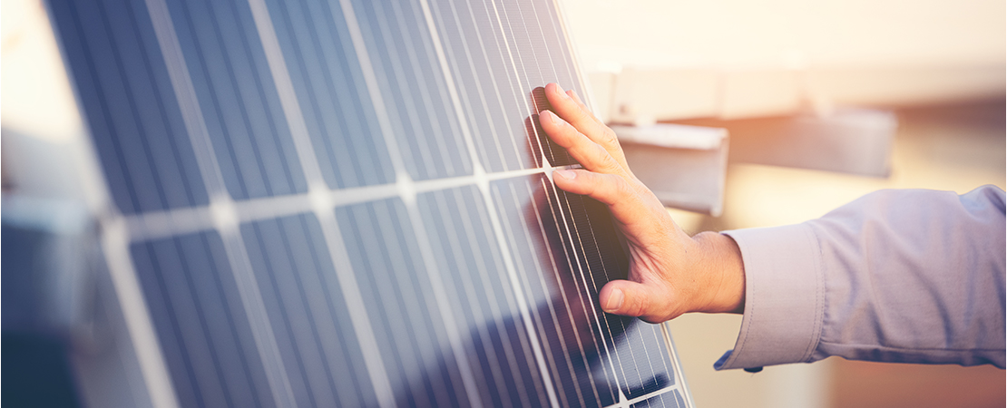 Hand einer Person, die eine Photovoltaik-Solaranlage bei Sonnenlicht berührt. Die glänzenden Solarmodule reflektieren das Licht und sind in Nahaufnahme zu sehen.