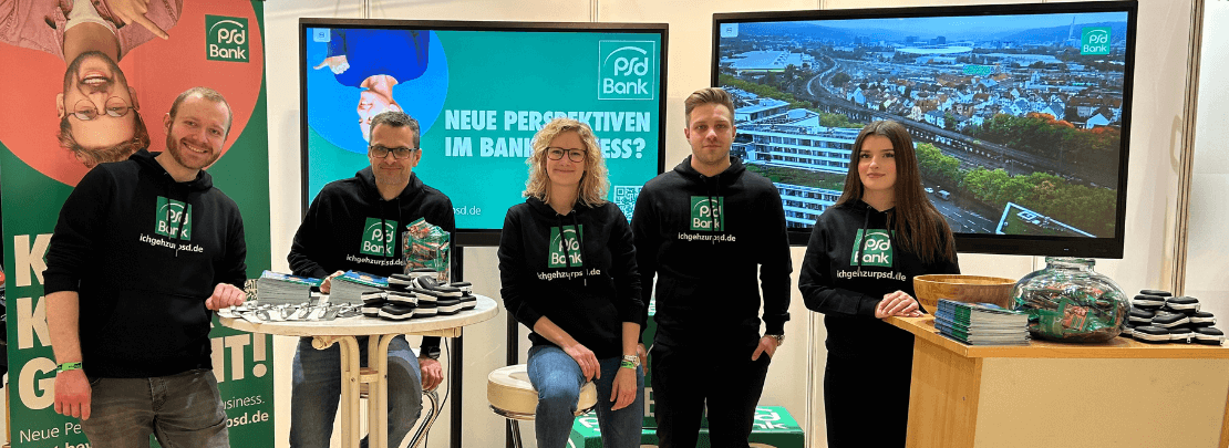 Fünf Mitarbeitende der PSD Bank stehen an einem Messestand mit Bildschirmen und Werbematerialien, alle tragen schwarze Hoodies mit dem Slogan „ichgehezurpsd.de“.