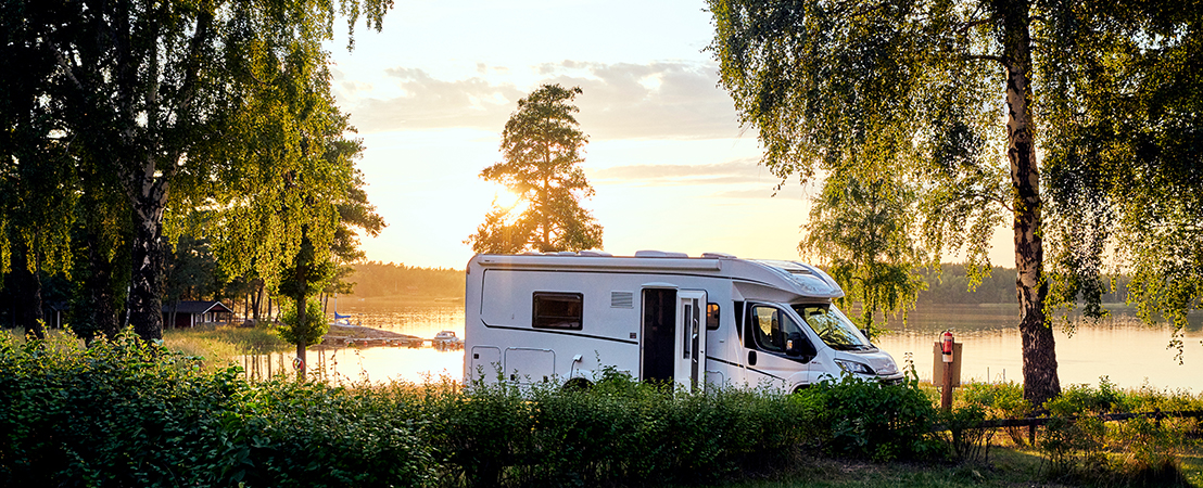 Wohnmobil steht bei Sonnenuntergang idyllisch an einem See zwischen Bäumen auf einem Naturcampingplatz. Symbolbild für Reisen mit Wohnmobil.