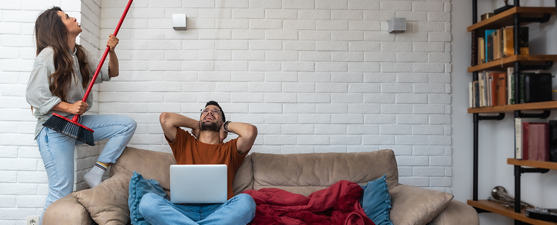 Eine Frau klopft mit einem Besen an die Decke, während ein Mann genervt auf dem Sofa sitzt – wegen Lärm von oben. Symbolbild für: Was tun bei lauten Nachbarn?