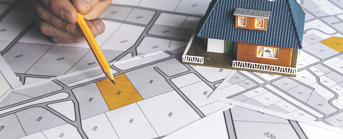 Hand zeigt mit Bleistift auf ein markiertes Grundstück auf einem Katasterplan, daneben steht ein Miniaturhaus – symbolisch für Grundstücksgrenzen und Bauplanung.