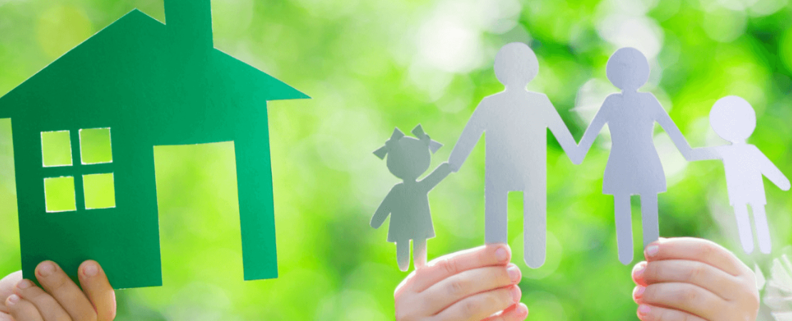 Hände halten eine grüne Haus-Silhouette und eine ausgeschnittene Papierfamilie mit zwei Erwachsenen und zwei Kindern vor einem sonnigen, grünen Hintergrund. Symbolbild für Familienglück, Eigenheim, nachhaltiges Wohnen und Immobilienfinanzierung.