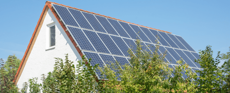 Einfamilienhaus mit Photovoltaikanlage auf dem Dach, umgeben von grünen Bäumen, unter klarem blauem Himmel. Die Solarpaneele bedecken die gesamte Dachfläche und stehen für nachhaltige Energiegewinnung. Symbolbild für erneuerbare Energien, Solarstrom und umweltfreundliches Wohnen.