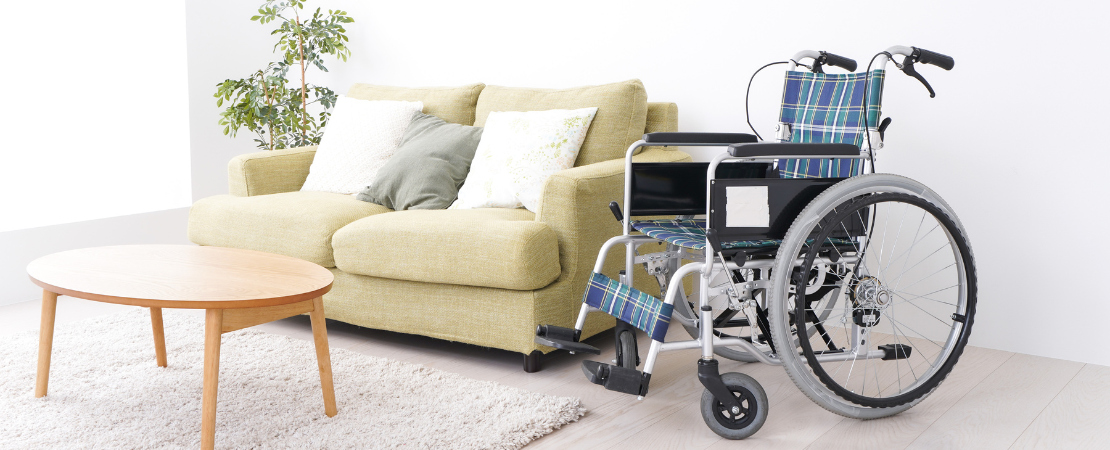 Rollstuhl mit blau-kariertem Sitzbezug steht in einem barrierefreien Wohnzimmer neben einem grünen Sofa mit dekorativen Kissen. Ein runder Holztisch und ein heller Teppich ergänzen die moderne, freundliche Einrichtung. Symbolbild für Inklusion, barrierefreies Wohnen und häusliche Pflege.