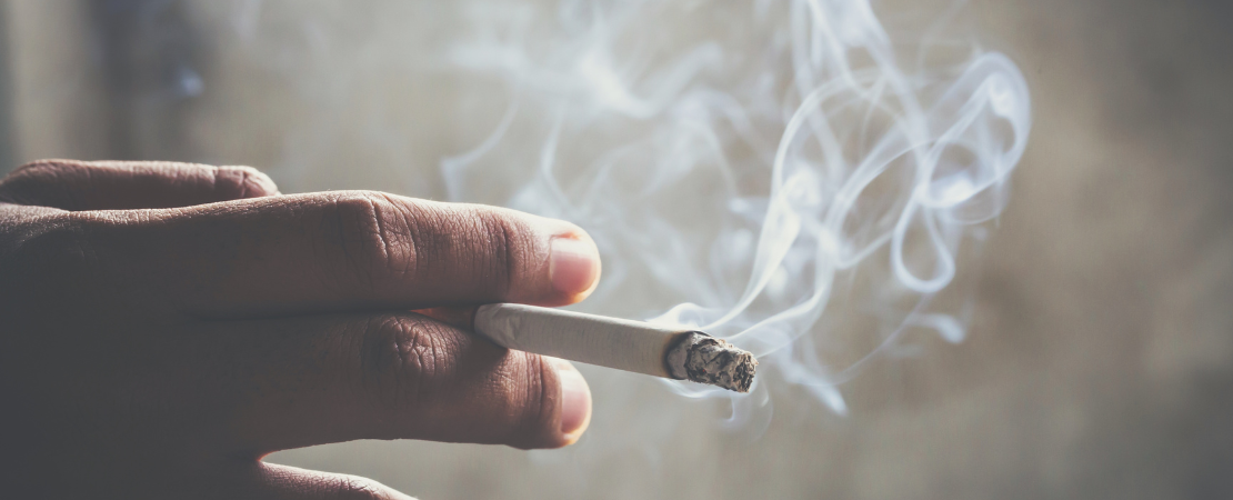 Nahaufnahme einer Hand, die eine brennende Zigarette hält, während Rauch in die Luft steigt. Der Hintergrund ist unscharf und gedämpft beleuchtet. Symbolbild für Rauchen, Tabakkonsum.