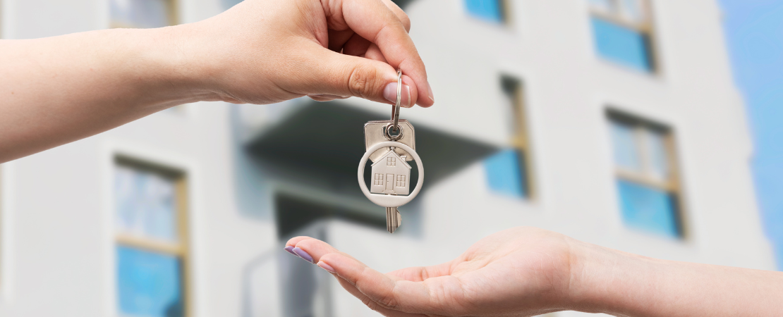Schlüsselübergabe vor einem modernen Wohngebäude: Eine Hand überreicht einen Schlüsselbund mit einem Haus-förmigen Anhänger an eine andere Hand. Unscharfer Hintergrund mit einem mehrstöckigen Apartmenthaus symbolisiert Mietbeginn oder Wohnungsübergabe.