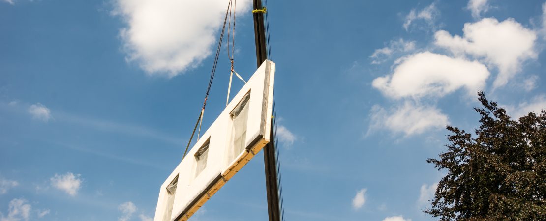 Ein Kran mit einer Fertighauswand am Seil, die herabgelassen wird.